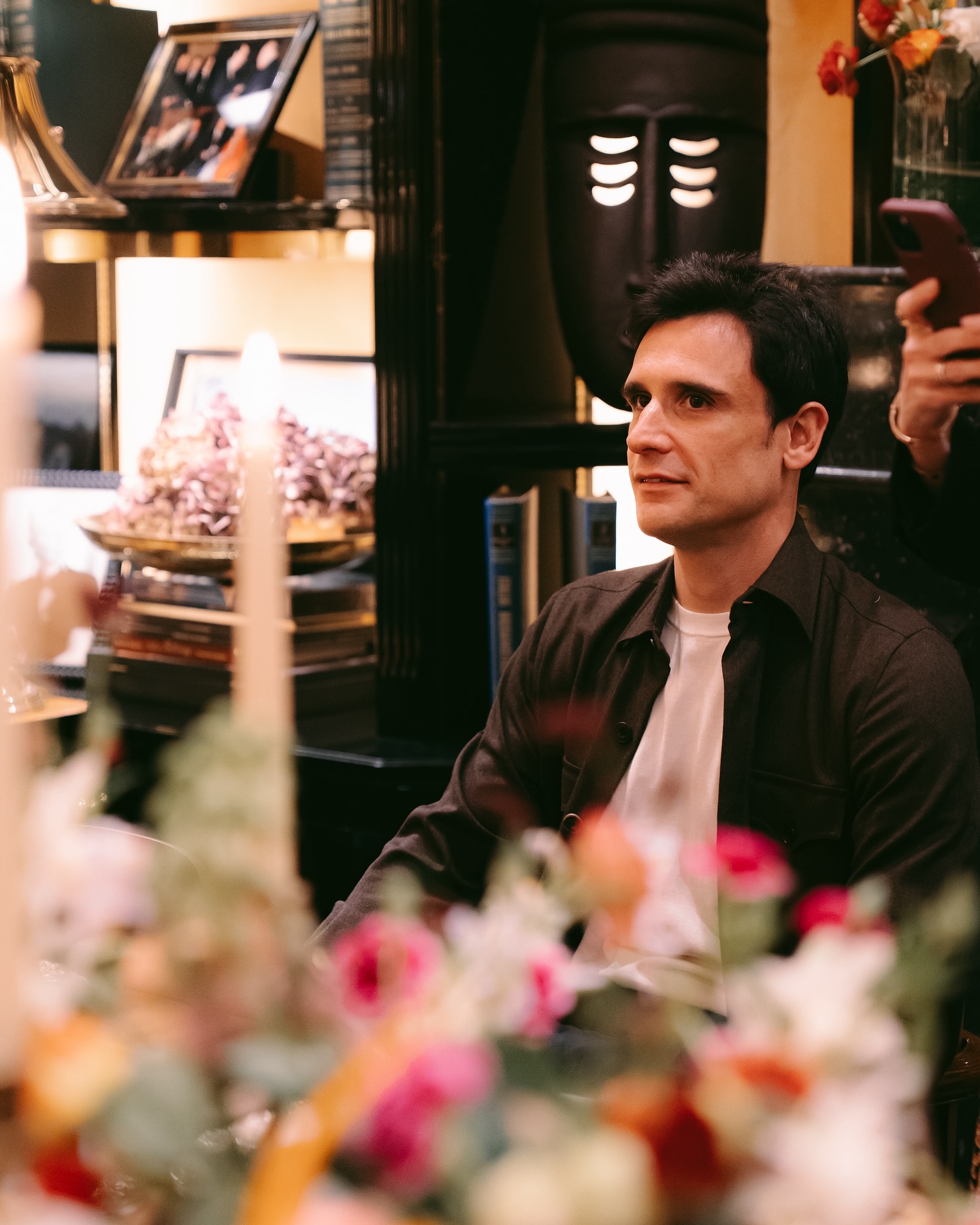 A man with dark hair, wearing a dark shirt over a white t-shirt, sits indoors near a table with flowers and candles. Behind him are books, framed photos, and a decorative mask on a shelf.