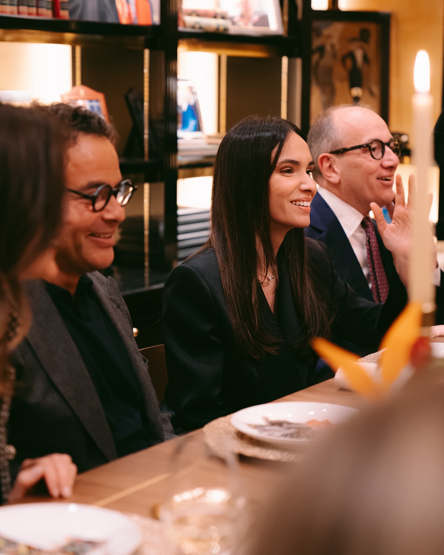 Three people in formal attire sit at a dinner table, smiling and engaging in conversation. There are plates and a candle on the table, with shelves and warm lighting in the background.