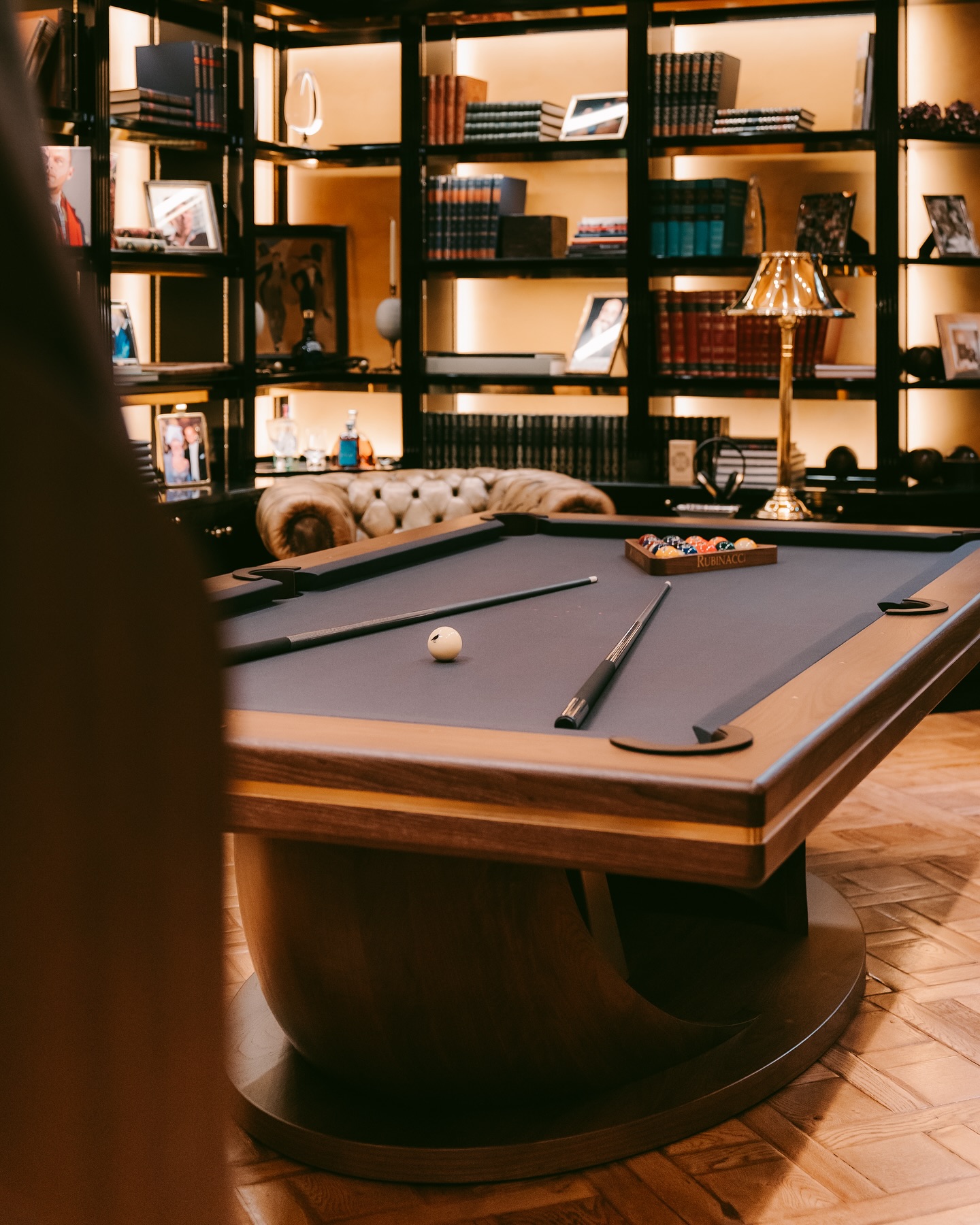 A stylish billiards table with cues and a triangle of balls sits in a cozy library room, surrounded by bookshelves filled with books and a tufted sofa in the background. Warm lighting creates an inviting atmosphere.
