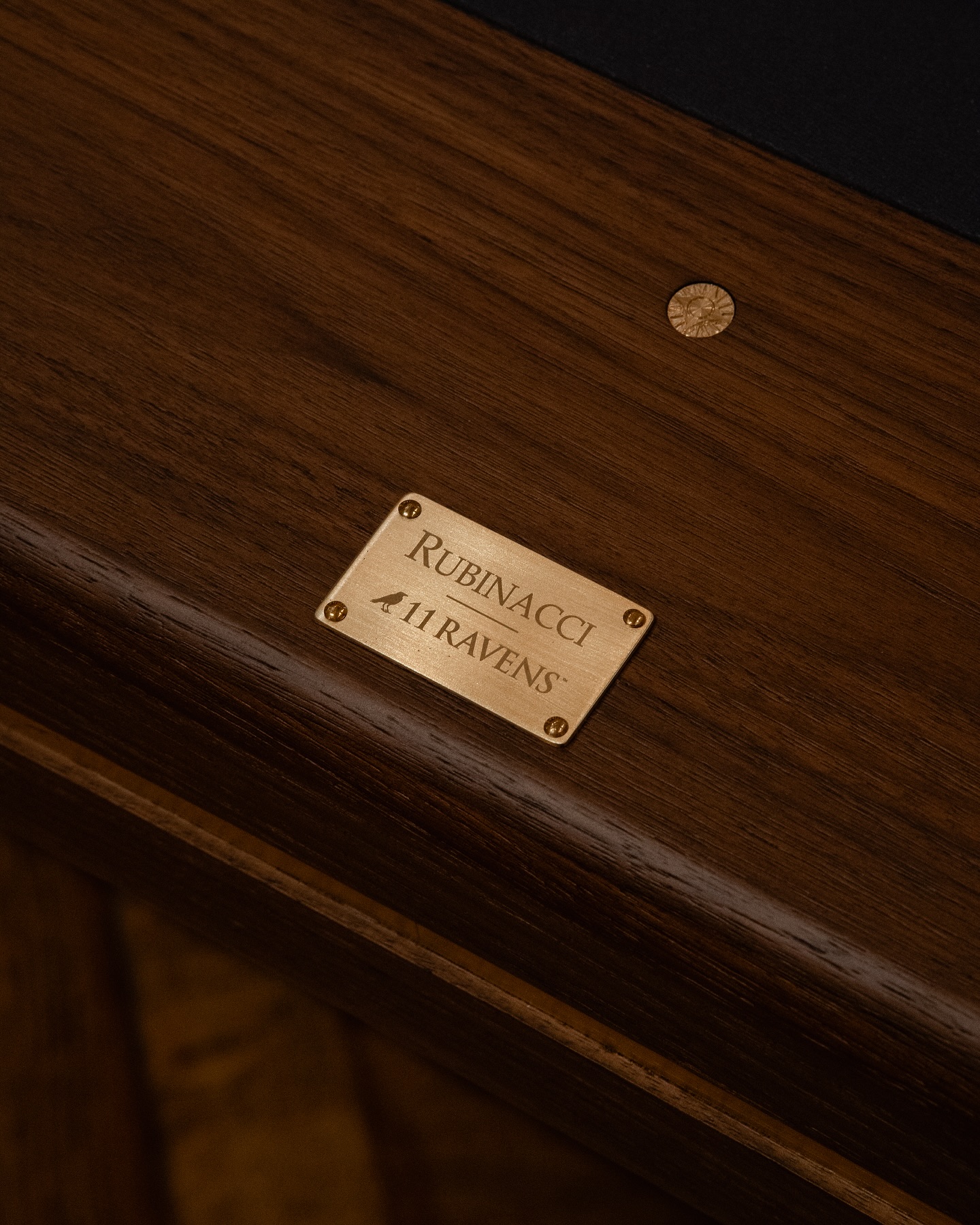 A brass nameplate engraved with Rubinacci x 11 Ravens is mounted on a polished dark wood surface. A decorative brass fastener is also visible nearby.