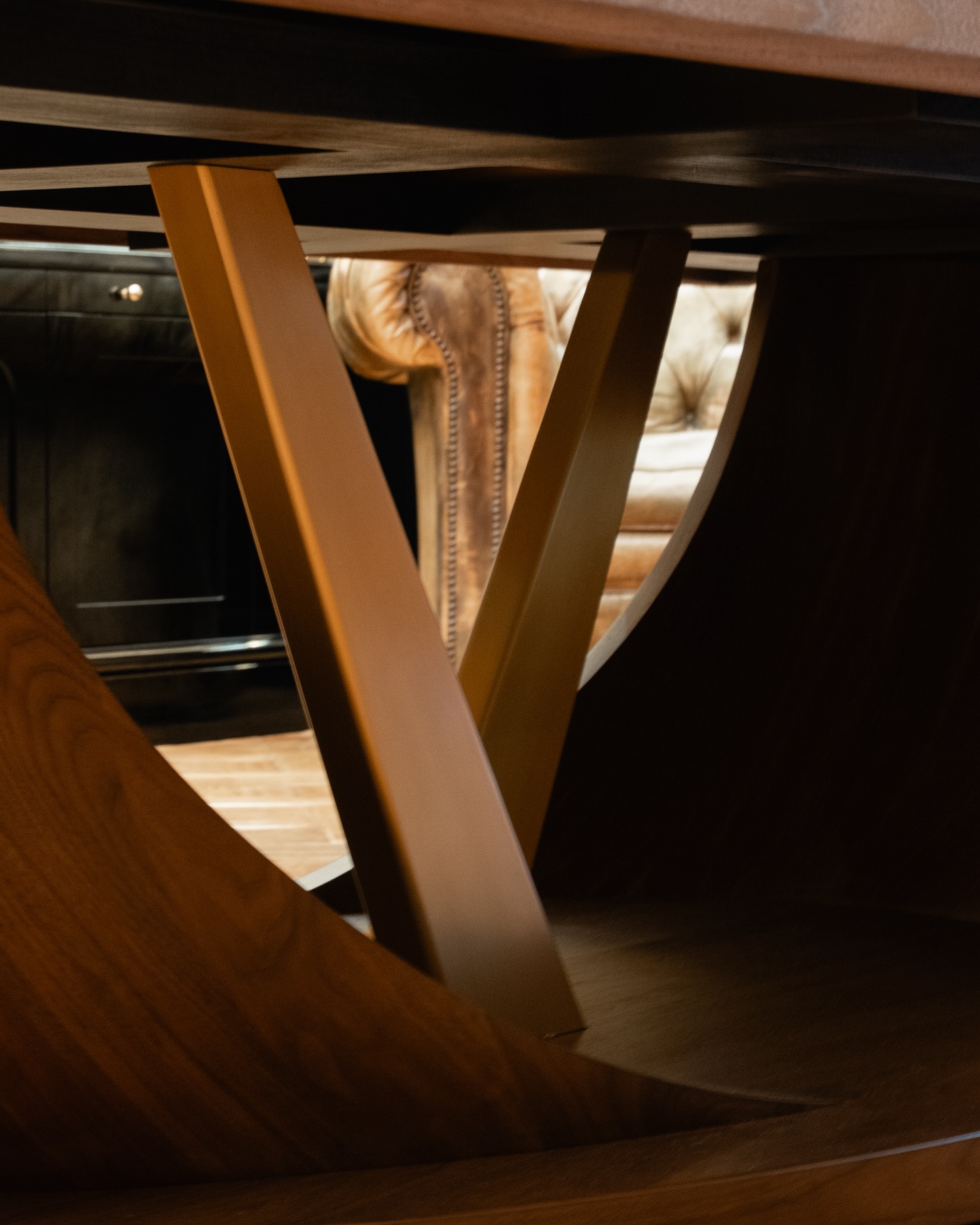 Close-up view of a wooden tables underside, showing intersecting angled supports, with part of a brown tufted leather armchair visible in the background. Warm lighting highlights the textures and materials.