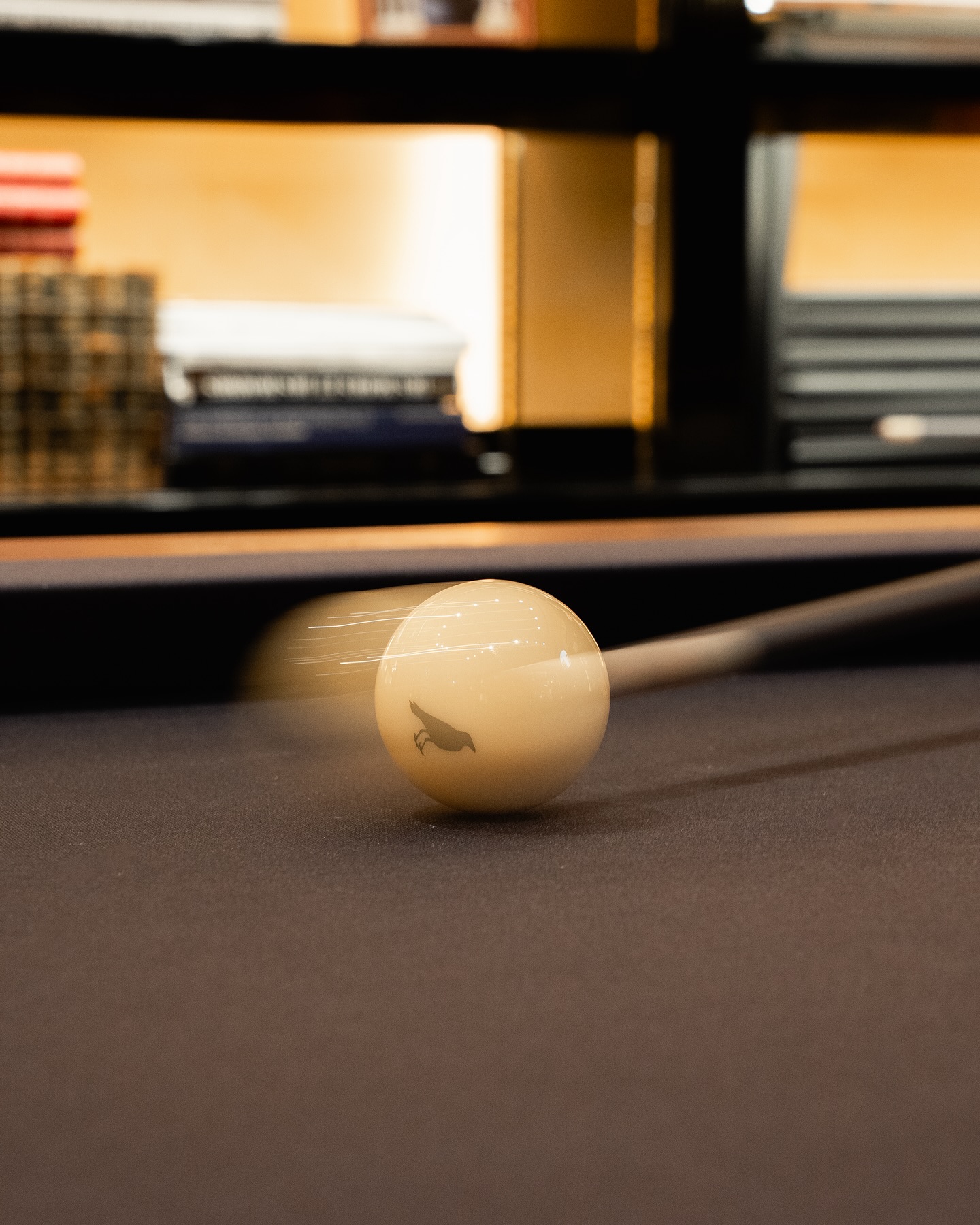 A close-up of a white billiard ball being struck by a cue stick, captured in motion on a pool table with blurred bookshelves in the background.