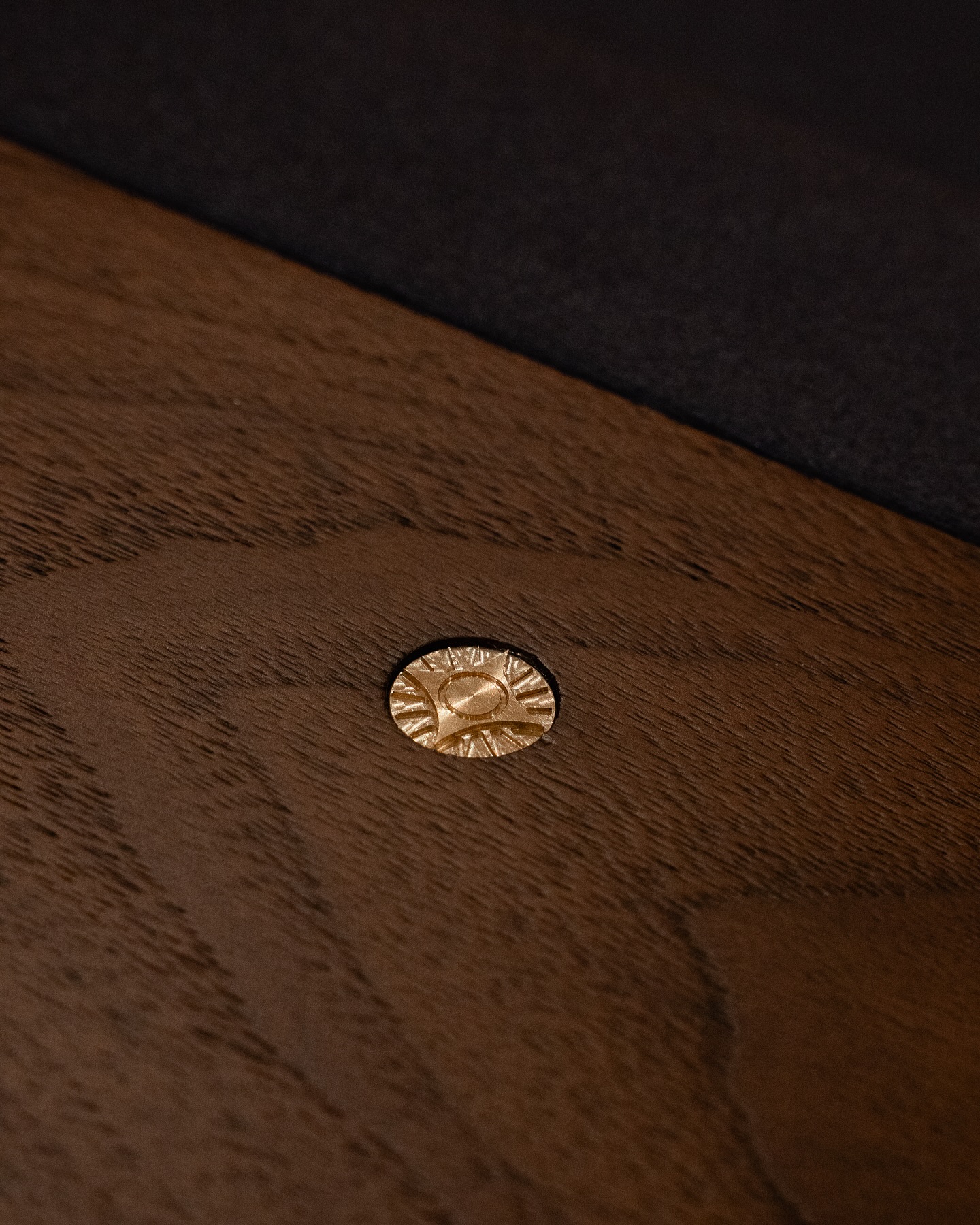 A close-up of a wooden surface with a decorative brass or gold screw, featuring an engraved sunburst pattern, partially reflecting light. The wood has a smooth, rich brown grain.