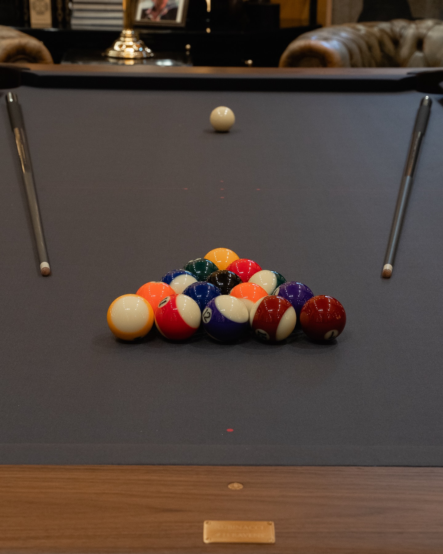A racked set of colorful billiard balls sits on a dark pool table, with two pool cues positioned on either side and the cue ball placed at the opposite end, ready for break.