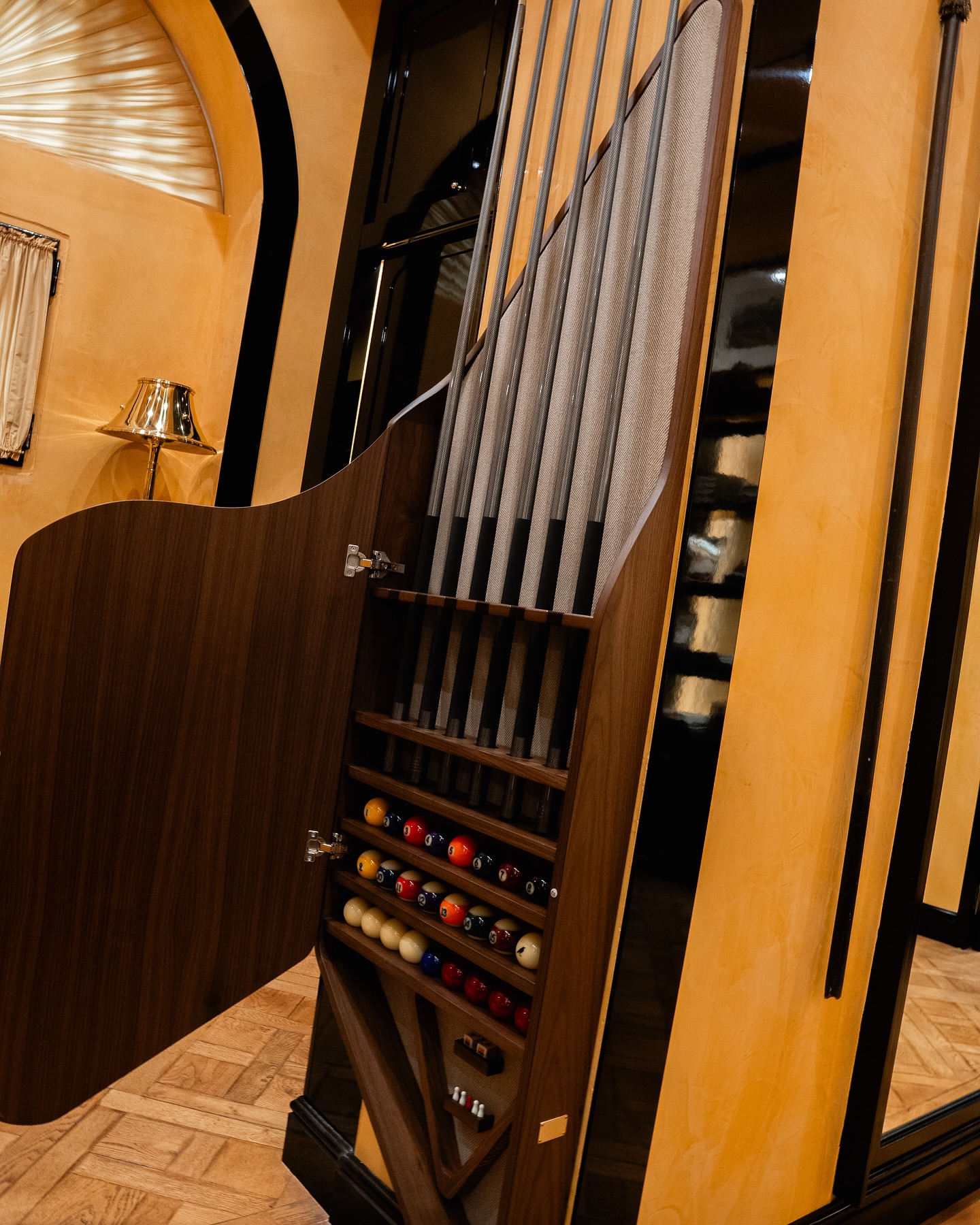 A stylish wooden cabinet with an open door reveals billiard cues and a neatly organized rack of pool balls inside, set against a warmly lit, elegant interior with tan and black walls.