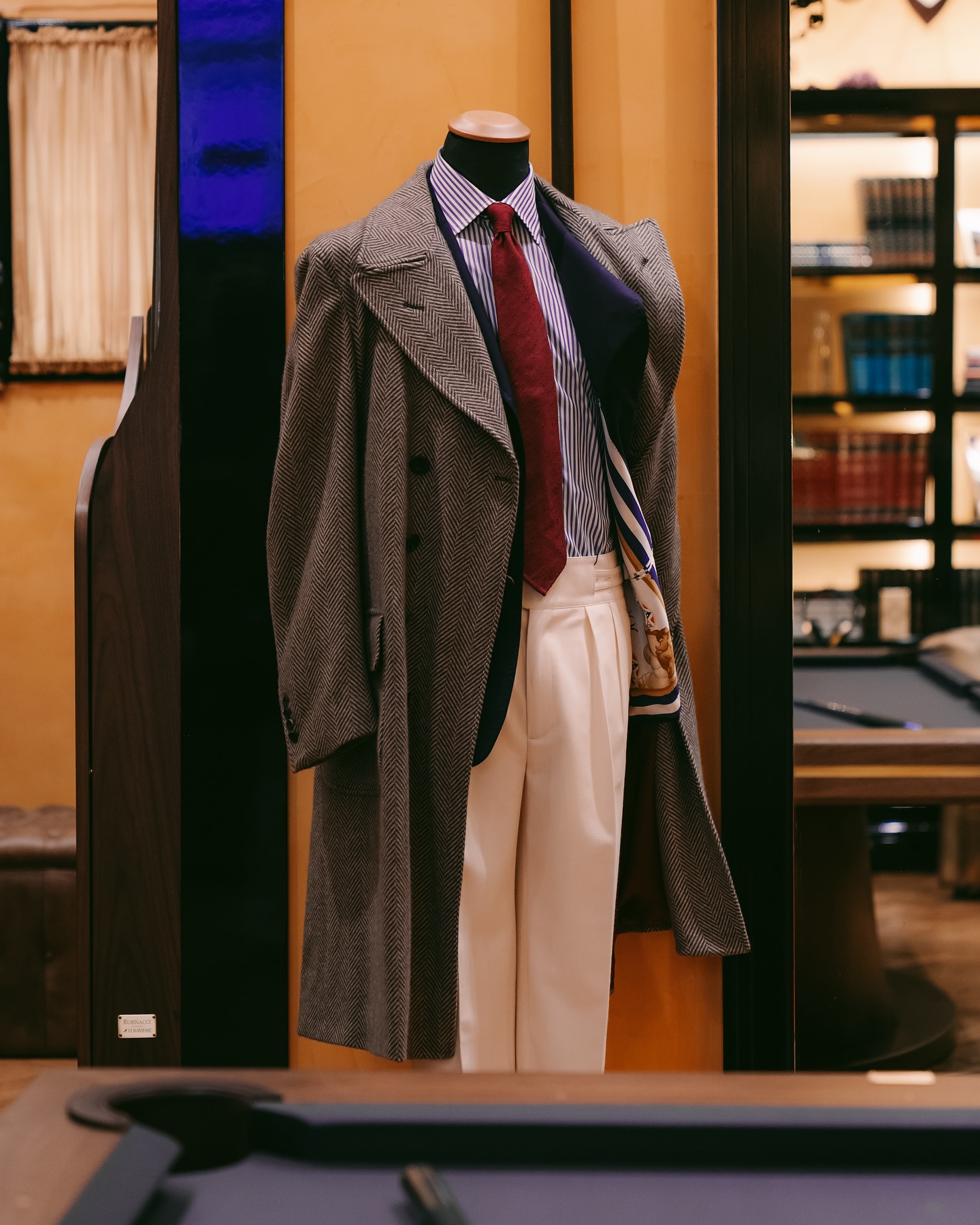 A headless mannequin dressed in a gray overcoat, blue and white striped shirt, red tie, white trousers, and a brown shoulder bag, stands in a stylish, warmly lit room with bookshelves in the background.