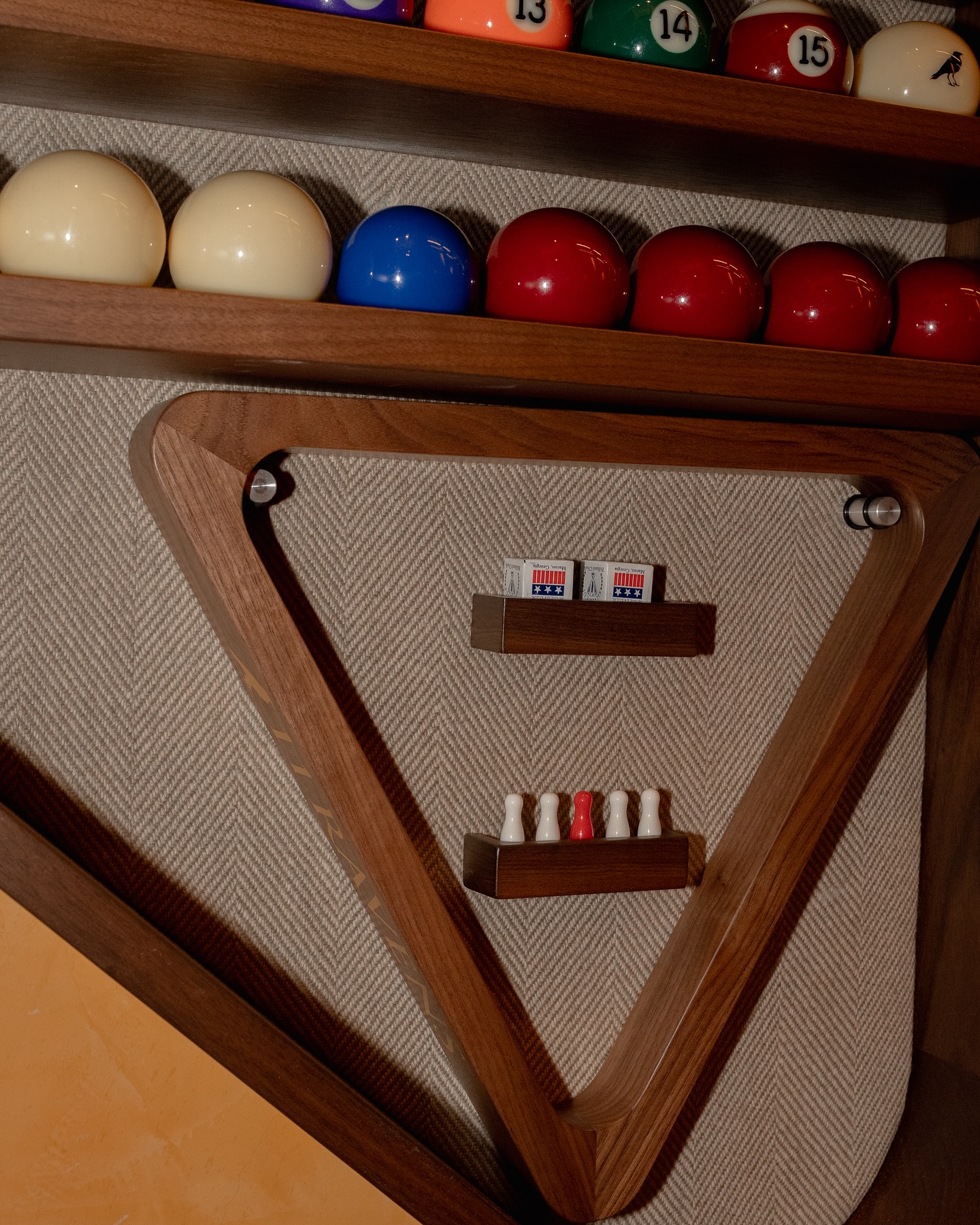A wooden wall shelf shaped like a triangle holds billiard balls on top, with two decks of playing cards and four red-and-white chess pawns displayed on small shelves inside the triangle.