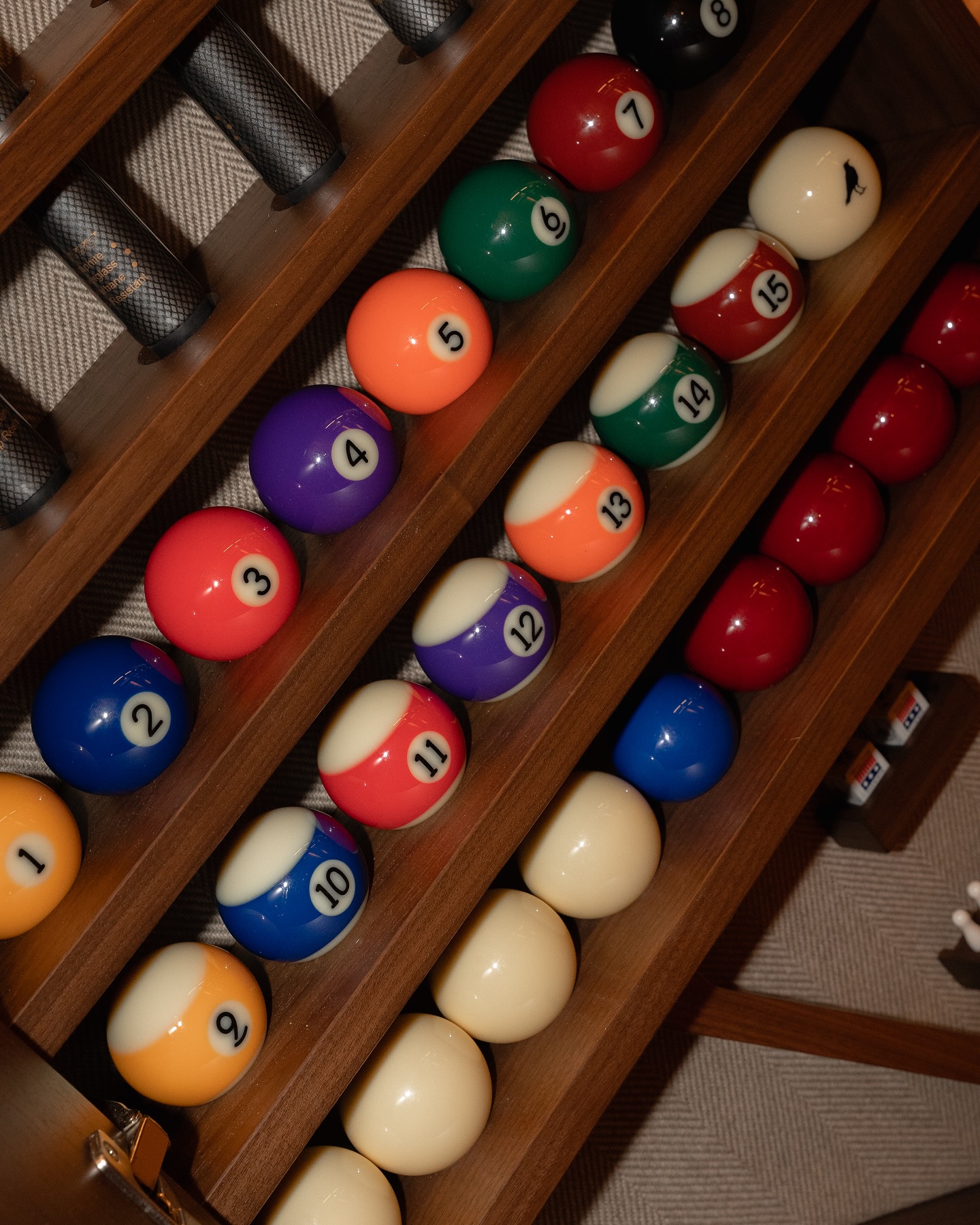 A wooden rack holds colorful billiard balls, each numbered, neatly arranged in rows. Cue sticks are stored above the balls, and part of a pool table is visible in the lower right corner.