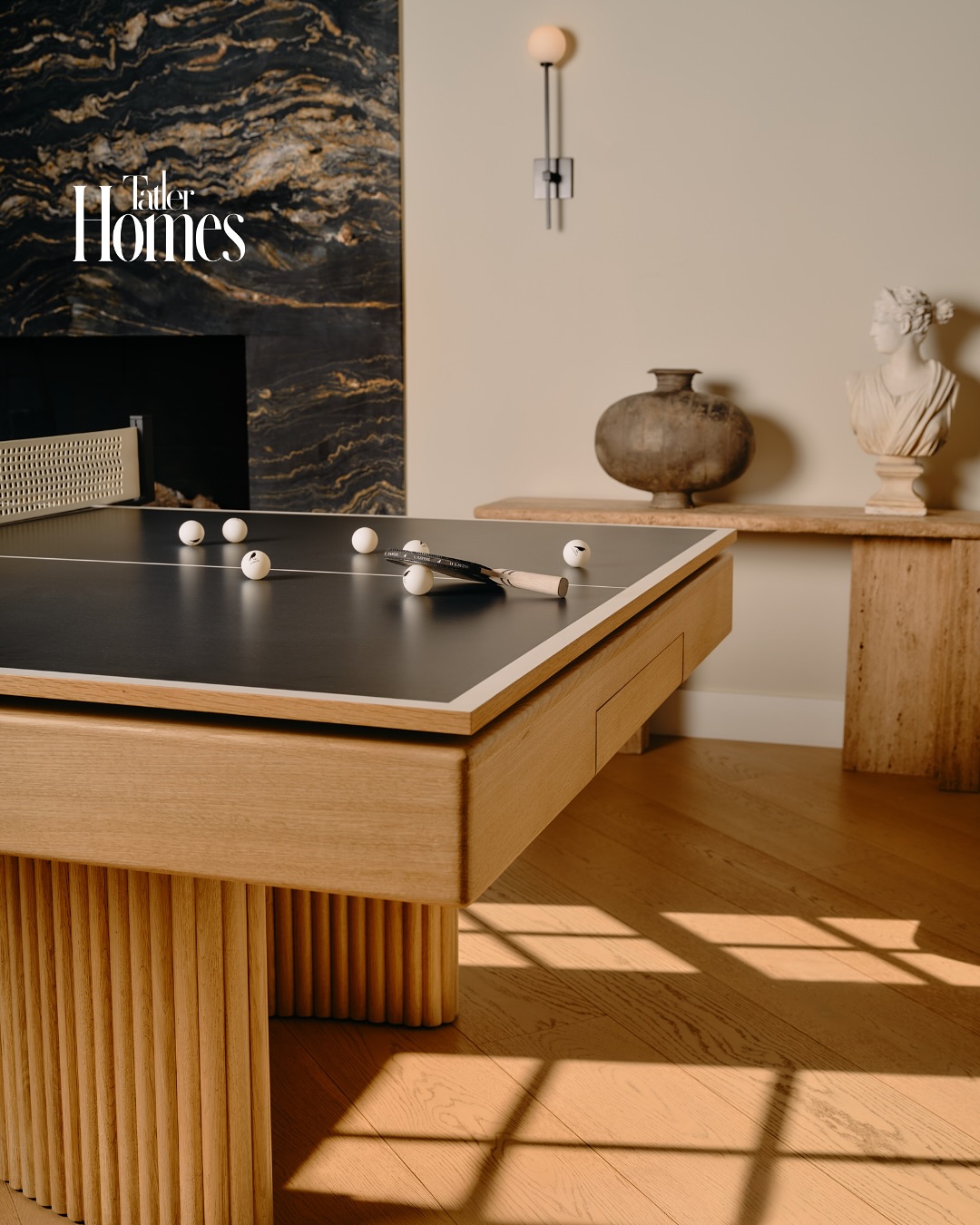 A modern room styled by an interior designer features a wooden ping pong table with white balls and paddles. In the background, a marble fireplace, a vase and bust-topped table, and sunlight highlight the elegant wooden floor.