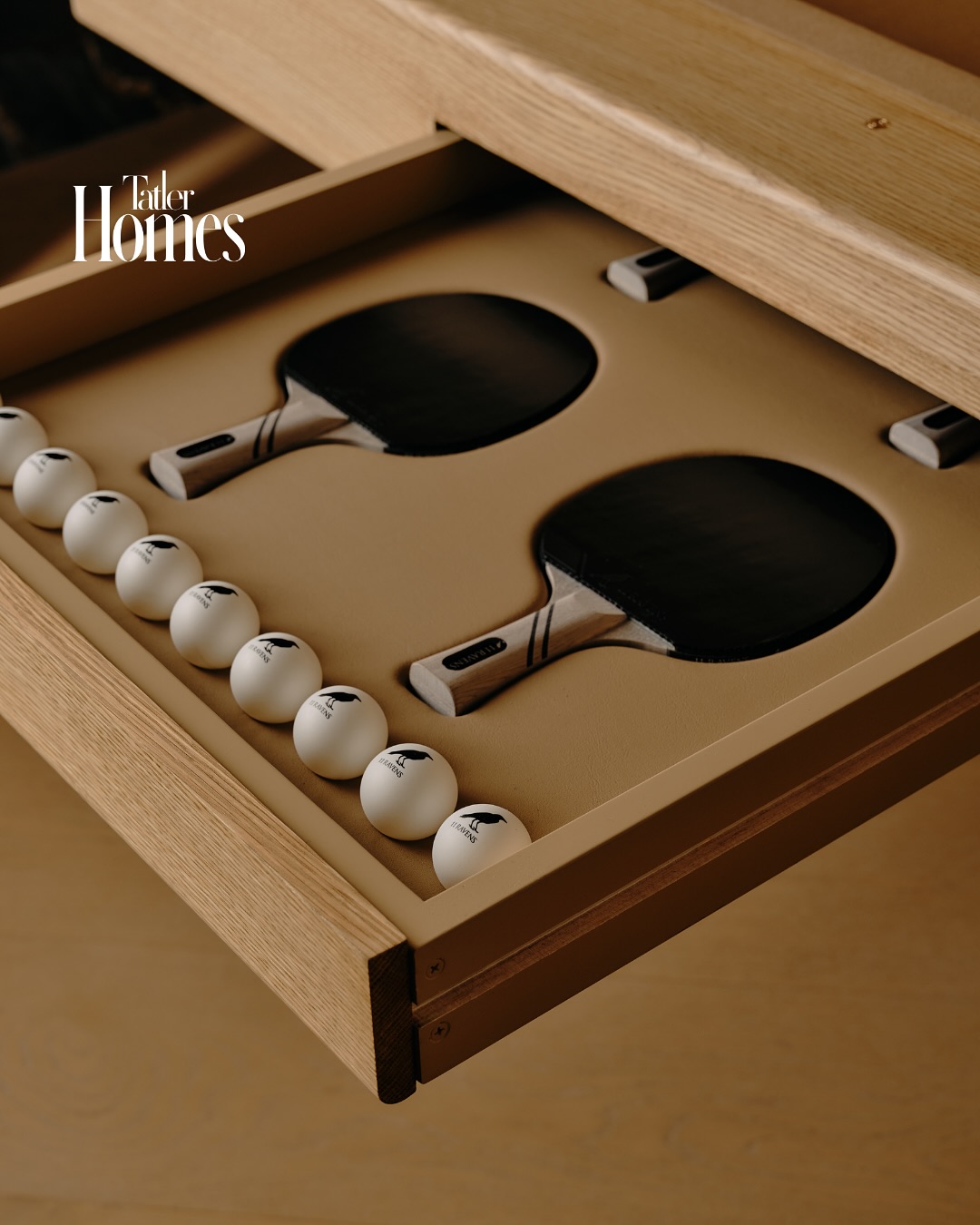 An open wooden drawer displays two black ping pong paddles and a row of white ping pong balls in a custom foam insert—an interior designer’s touch. Tatler Homes is written in the top left corner.