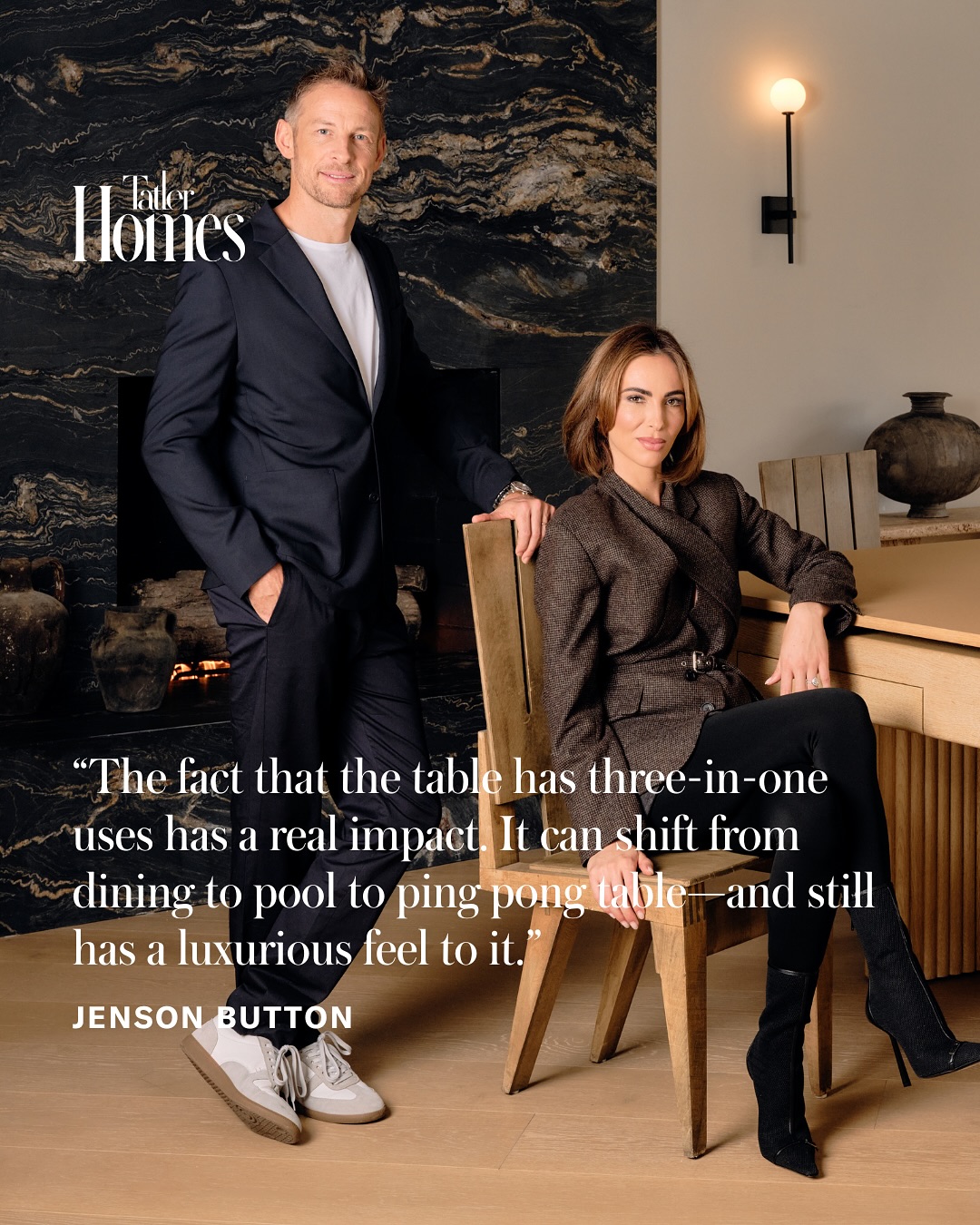 A man in a suit stands next to a woman sitting on a chair in a modern dining room with a large stone wall and wooden table. Text overlay features Formula One legend Jenson Buttons quote about this versatile table, admired by any interior designer.