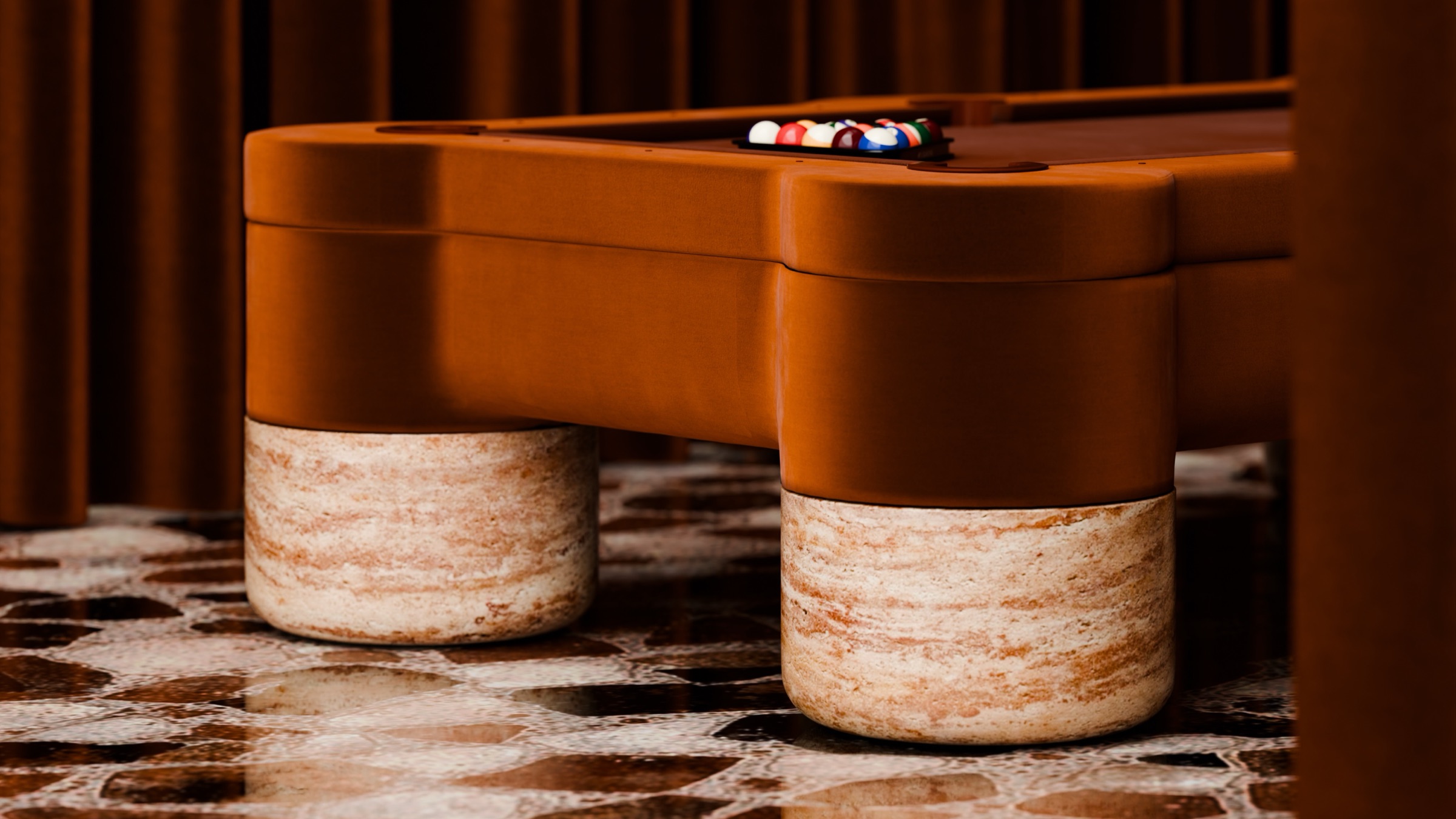 A close-up of the Marc Ange Pool Table highlights its brown finish and thick cylindrical stone legs on a patterned tile floor, with pool balls racked in a triangle at one end.