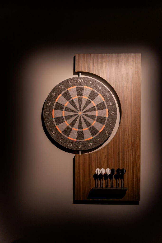 A dartboard mounted on a wooden backboard is illuminated on a wall, with six darts—three black and three white—placed on a holder below the board, showcasing a Recent Work - 11 Ravens design.