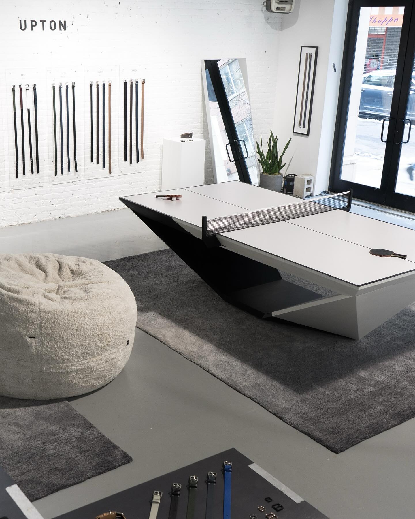 A modern showroom with a sleek white ping pong table, a large beige bean bag chair, gray rugs, a wall display of belts, a leaning mirror, a plant, and large windows letting in natural light.