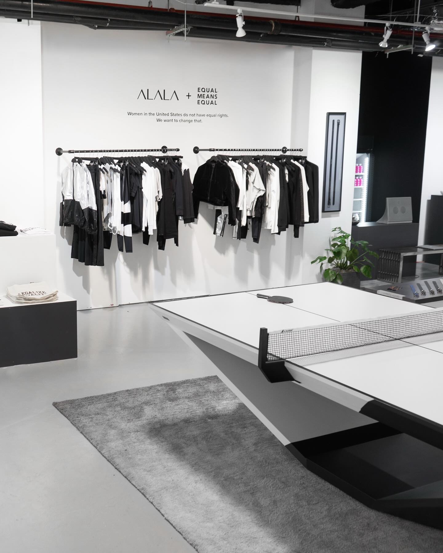 A modern store interior features a clothing rack with black and white garments, a ping pong table in the foreground, and a wall sign reading “ALALA + Equal Means Equal.” The space has minimalist decor and gray flooring.