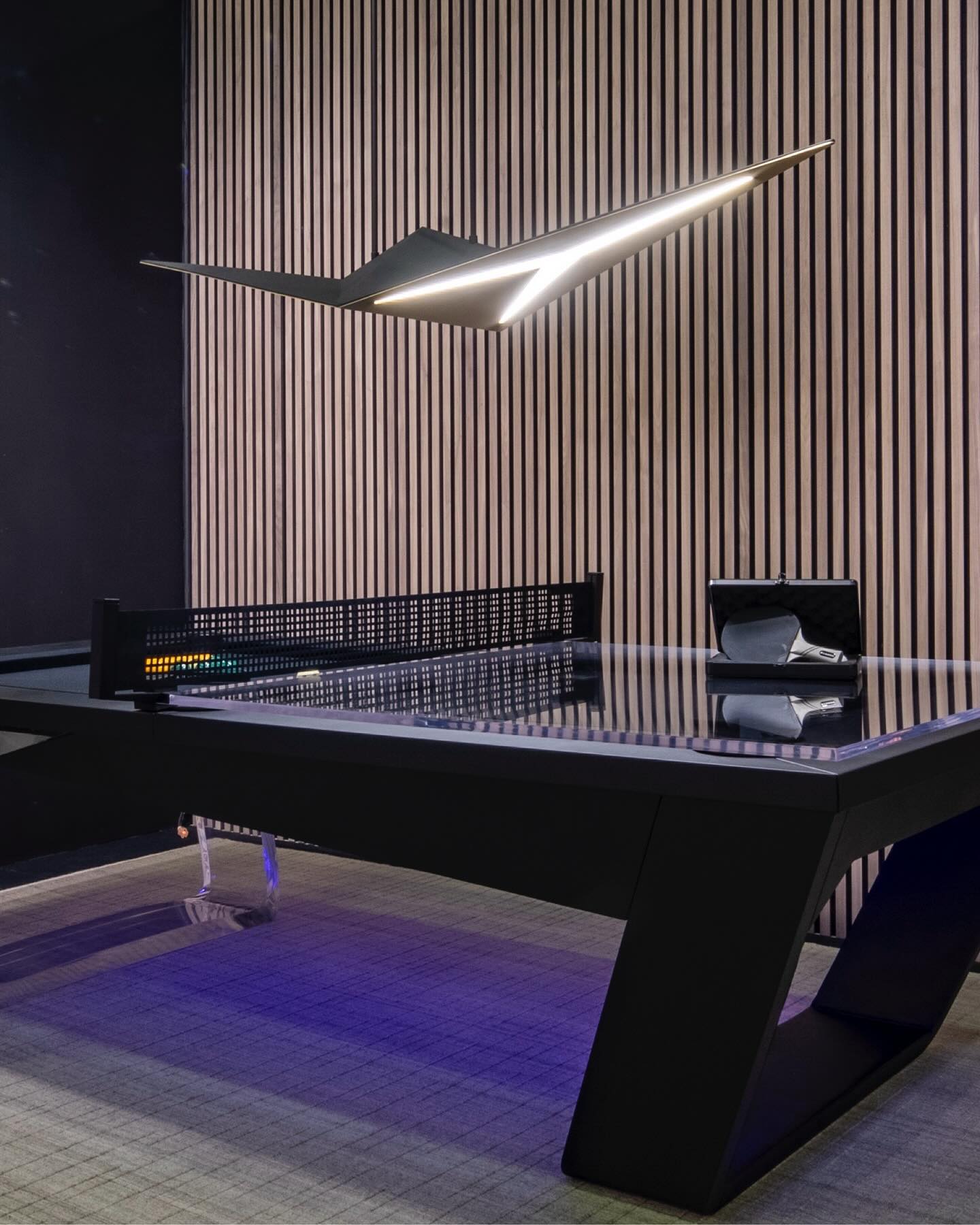 A modern black ping pong table with angular legs sits in front of a slatted wood wall, illuminated by a geometric, wing-shaped ceiling light and purple floor lighting.