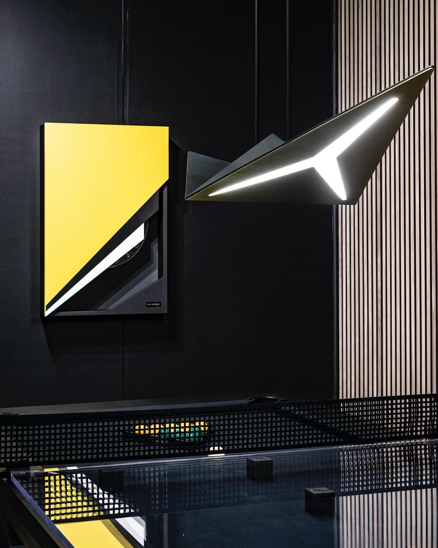 A modern interior features a geometric yellow, black, and white wall art piece beside a sculptural, angular light fixture emitting bright white light. The foreground includes part of a glossy glass table and a black mesh divider.