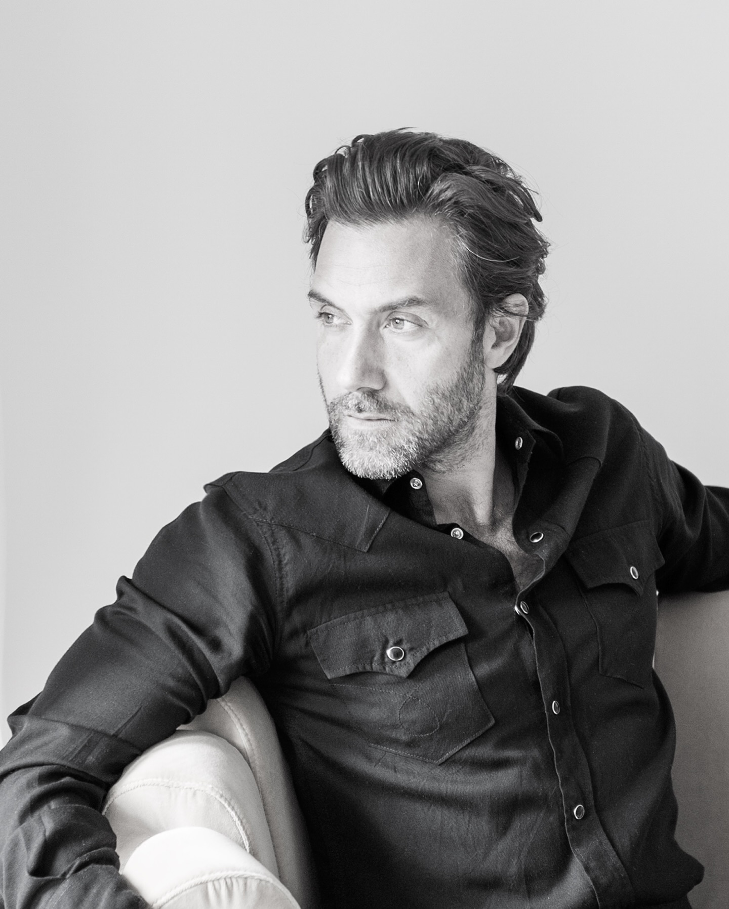 A man with wavy hair and a beard, wearing a dark button-up shirt, sits on a light-colored chair, looking to his left with a thoughtful expression in a bright, minimalist setting.