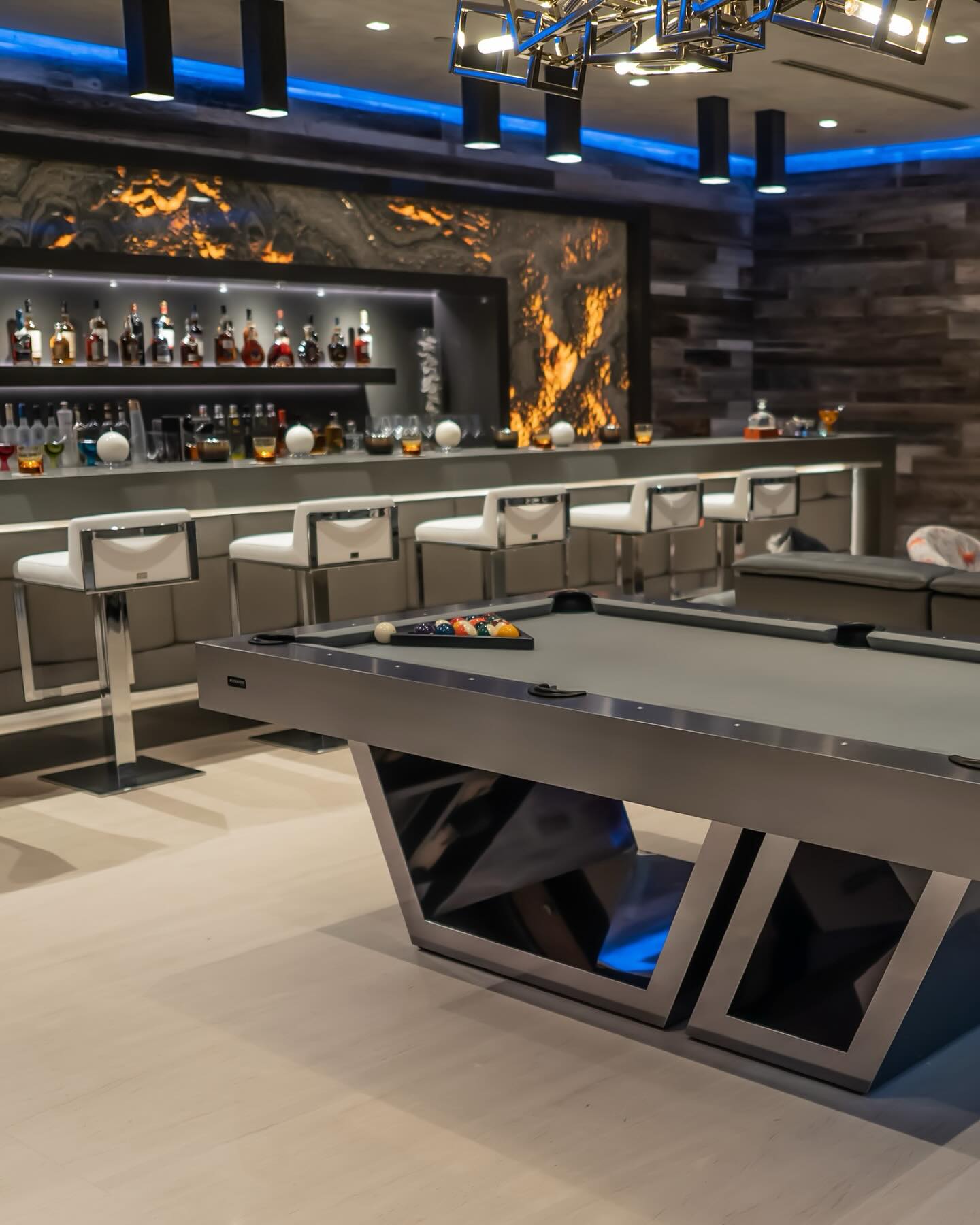 Modern game room with a sleek pool table in the foreground, and a contemporary bar area with high white chairs and a variety of liquor bottles in the background, under blue accent lighting.