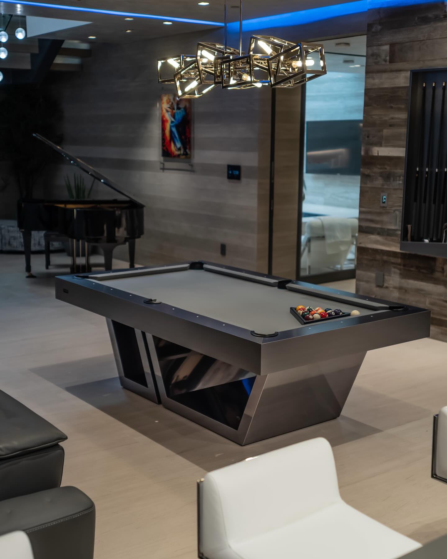 A modern room with a sleek black pool table, billiard balls racked, white furniture, a grand piano in the background, wood-paneled walls, and a geometric chandelier hanging above the table.