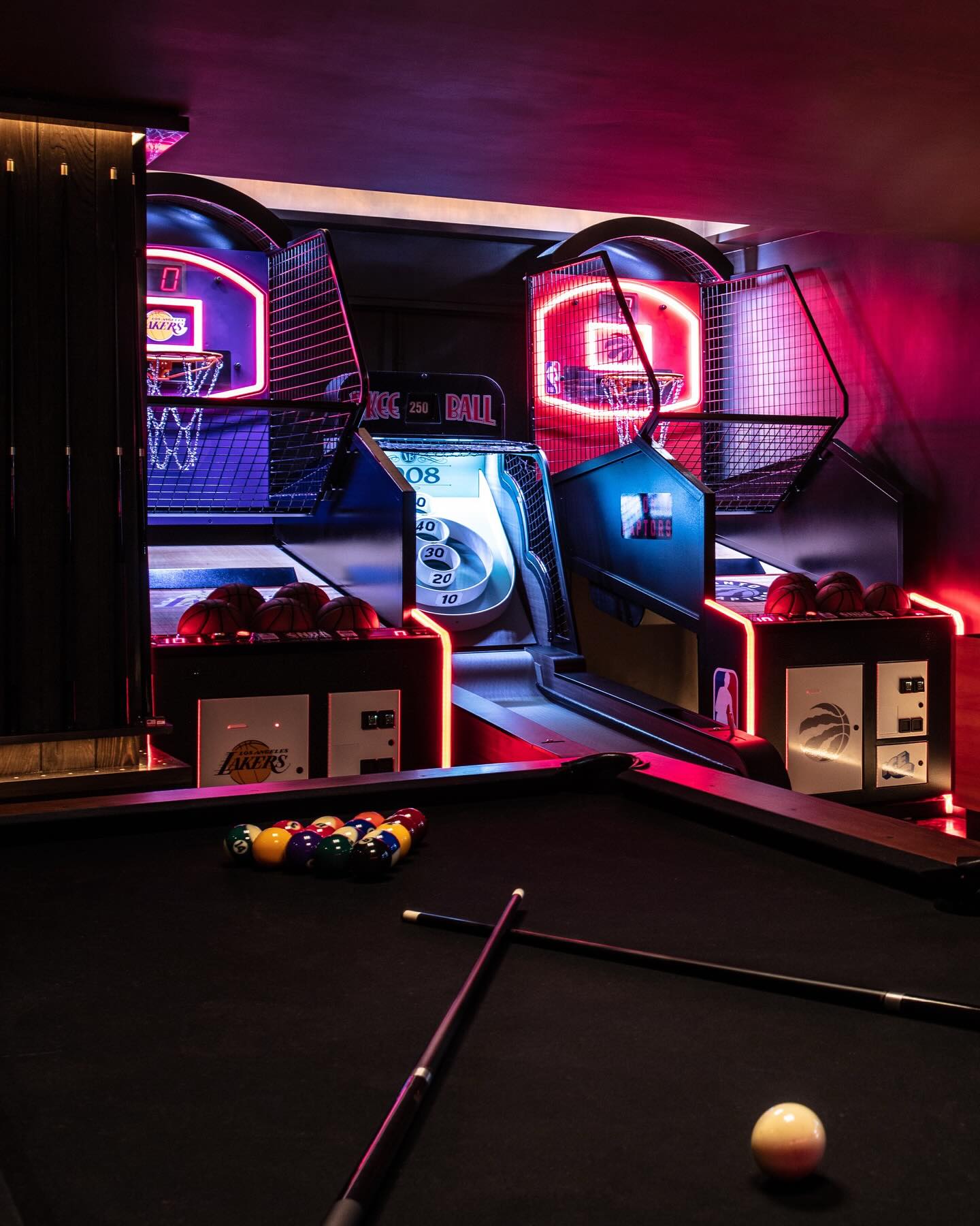 A dimly lit game room with a black pool table in the foreground and two basketball arcade machines with neon lights in the background. Pool balls and cues are on the table.