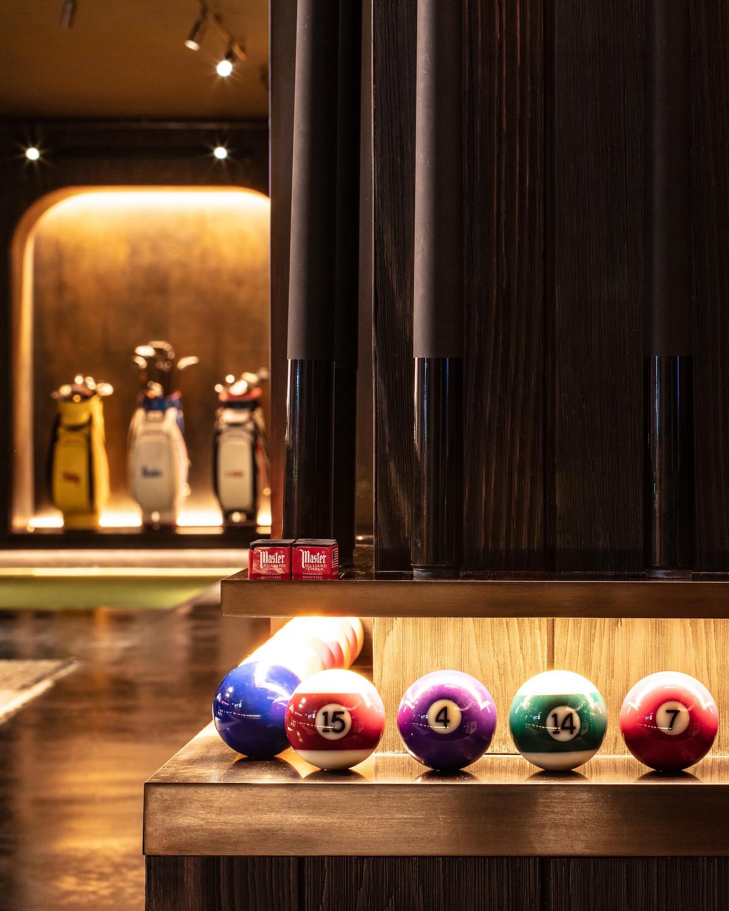 A rack of colorful pool balls, including the 15, 4, and 14 balls, sits on a wooden shelf. Pool cues stand upright nearby. In the background, golf bags are displayed in a softly lit area.