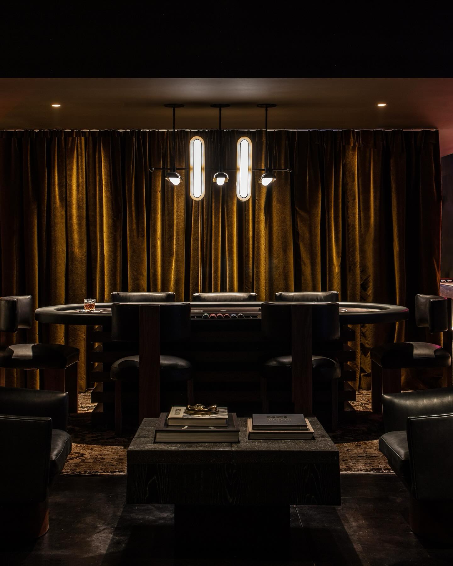 A dimly lit, luxurious room features a poker table surrounded by black leather chairs, a black coffee table with books, and gold velvet curtains. A modern chandelier hangs above, casting a warm, intimate glow.