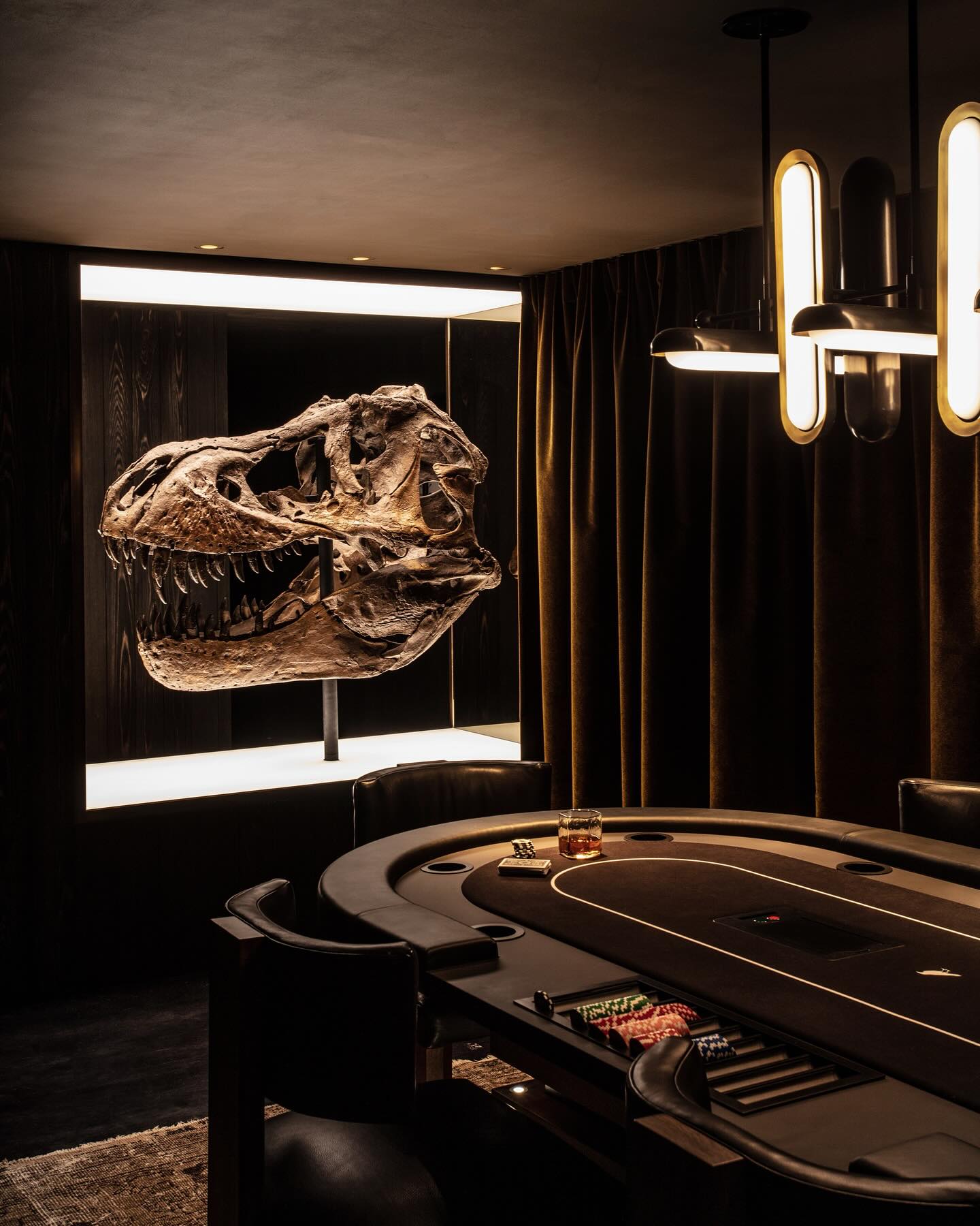 A large dinosaur skull is displayed in a lit wall niche behind a modern poker table with cards, chips, and a glass of whiskey. The room has dark walls, velvet curtains, and contemporary lighting.