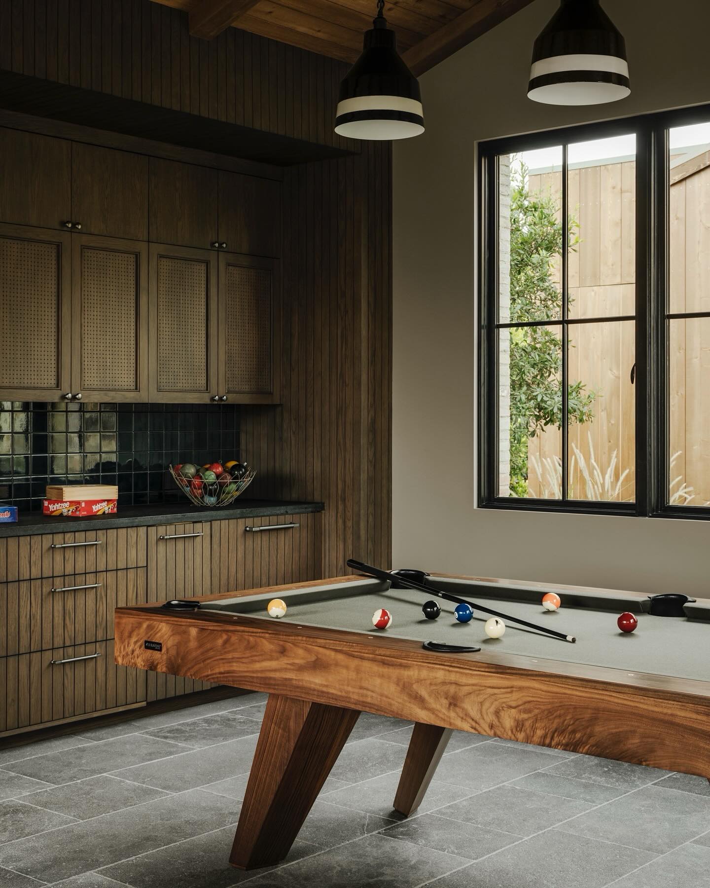 A modern game room with a wooden pool table holding balls and cues, a counter with games and a bowl of pool balls, wood cabinetry, and a large window letting in natural light.