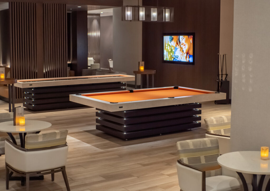 A modern lounge with two orange pool tables by Recent Work - 11 Ravens, white chairs, round tables, dim lamp lighting, and a wall-mounted TV displaying a movie scene. The space features wood accents and a relaxed ambiance.