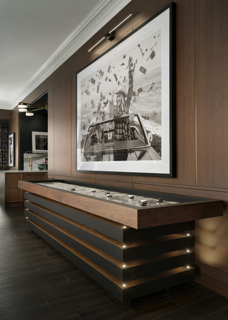 A modern game room with dark wood walls and floors features a sleek Recent Work - 11 Ravens shuffleboard table and a large black-and-white artwork of a person in a car with money flying in the air above them.