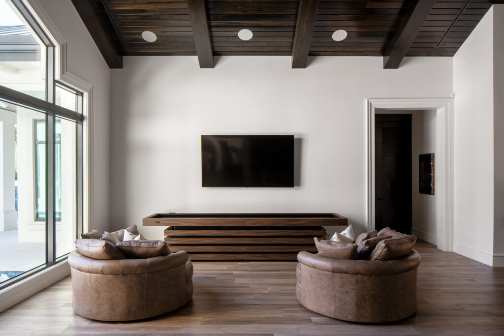 A modern living room with two round brown leather chairs, a large flat-screen TV mounted on a white wall, a wooden media console, wood floors, and exposed ceiling beams. Large windows let in natural light—see this Recent Work - 11 Ravens.