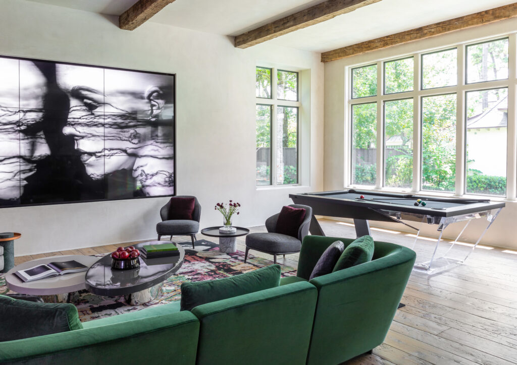A modern living room with large windows, a green sofa, round chairs, a Recent Work - 11 Ravens pool table, a colorful rug, and a wall-mounted TV displaying abstract black-and-white art. Natural light fills the spacious room.