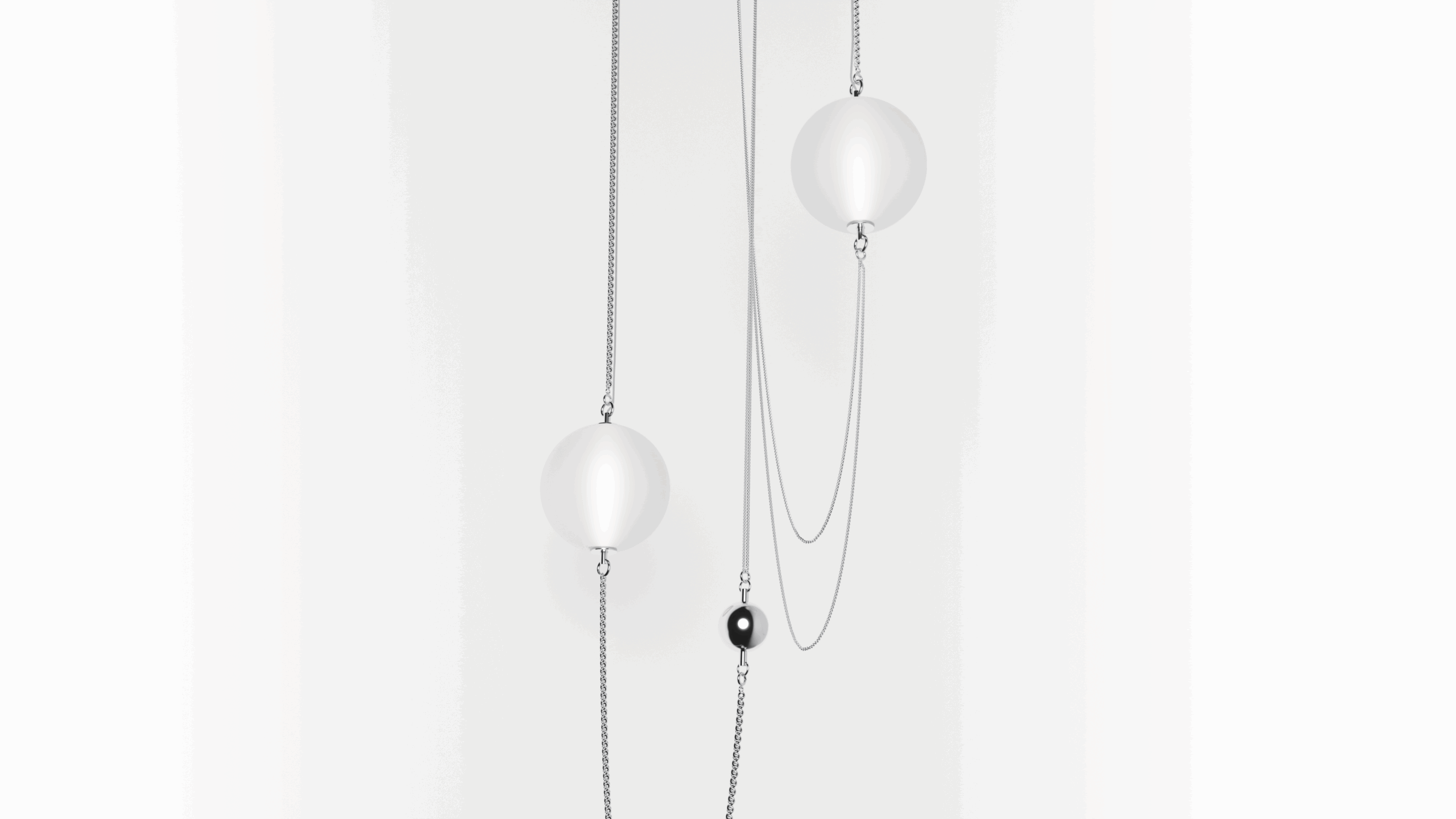 Two delicate silver chains hang vertically, each holding a smooth white sphere. A smaller black bead is suspended between them on one of the chains, all set against a plain white background.