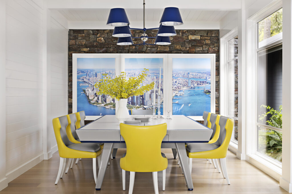 Bright dining room with a modern white table, yellow chairs, blue light fixture, and a vase of yellow flowers. Large windows reveal city and water views, with a stone accent wall—showcasing Recent Work - 11 Ravens’ refined design.