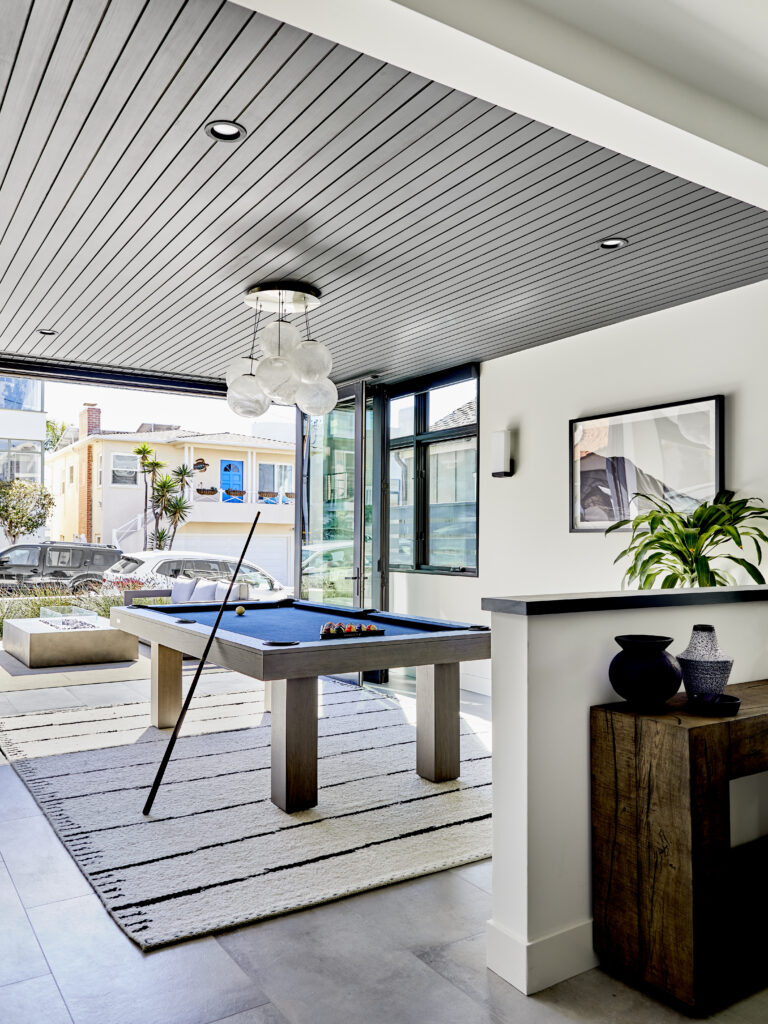 A modern, open room featuring a blue pool table by 11 Ravens on a striped rug, glass doors leading outside, a unique ceiling light, potted plants, and contemporary decor. Natural light fills this stylish, airy space—showcasing recent work.
