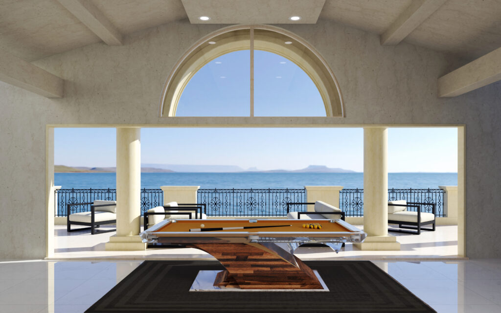A modern room featuring a unique wooden pool table by Recent Work - 11 Ravens rests on a dark rug, with large arched windows, white columns, and patio seating overlooking the ocean and distant hills under a bright blue sky.