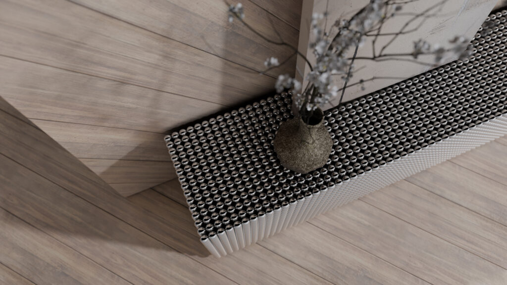 A textured metallic bench or radiator with a perforated pattern sits against a light wood-paneled wall, holding a brown vase with thin branches and delicate white flowers. The floor is also made of light wood.