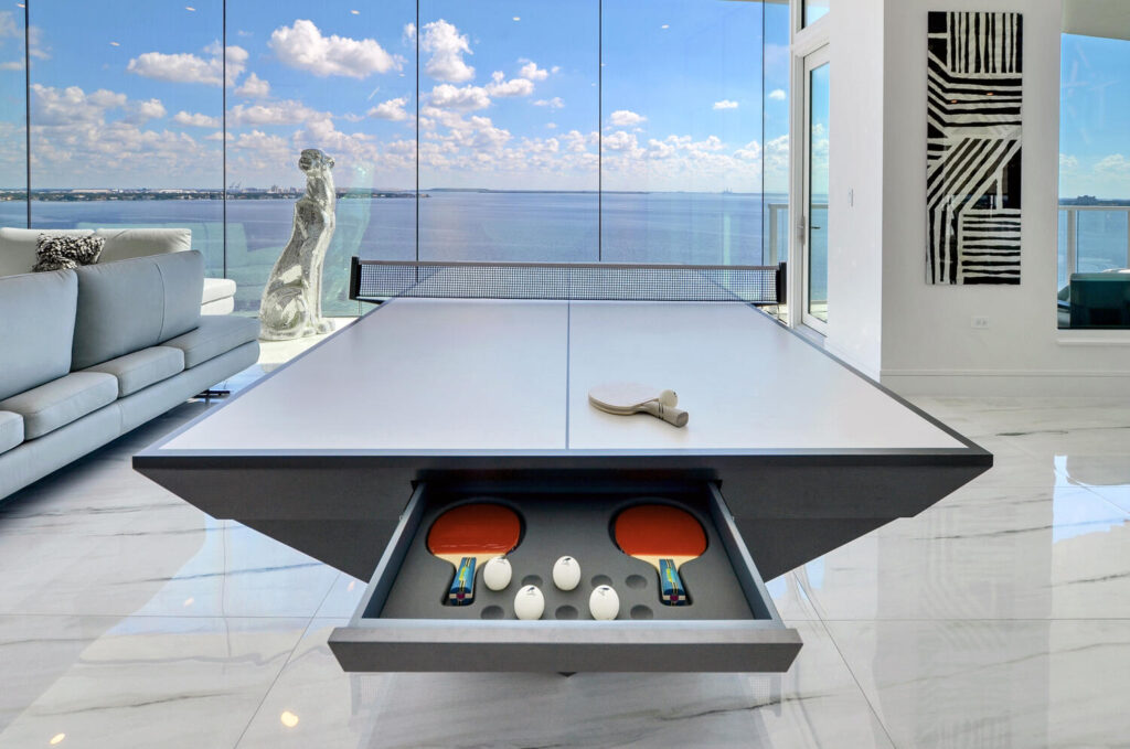 A modern table tennis setup by 11 Ravens in a bright room with a large glass wall overlooking water. The table’s drawer is open, revealing paddles and balls. Outside, a statue stands, while abstract art adorns the wall—recent work at its finest.