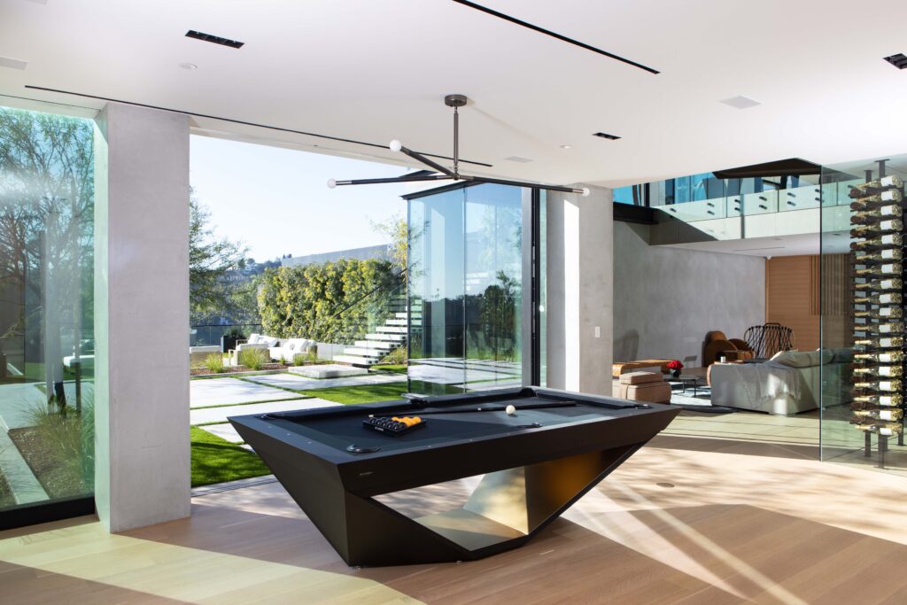 A modern living space featuring a sleek black pool table by 11 Ravens, large glass windows, and a wine display. Sunlight fills the room, offering views of a green lawn and outdoor stairs leading to a garden&mdash;another stunning recent work.
