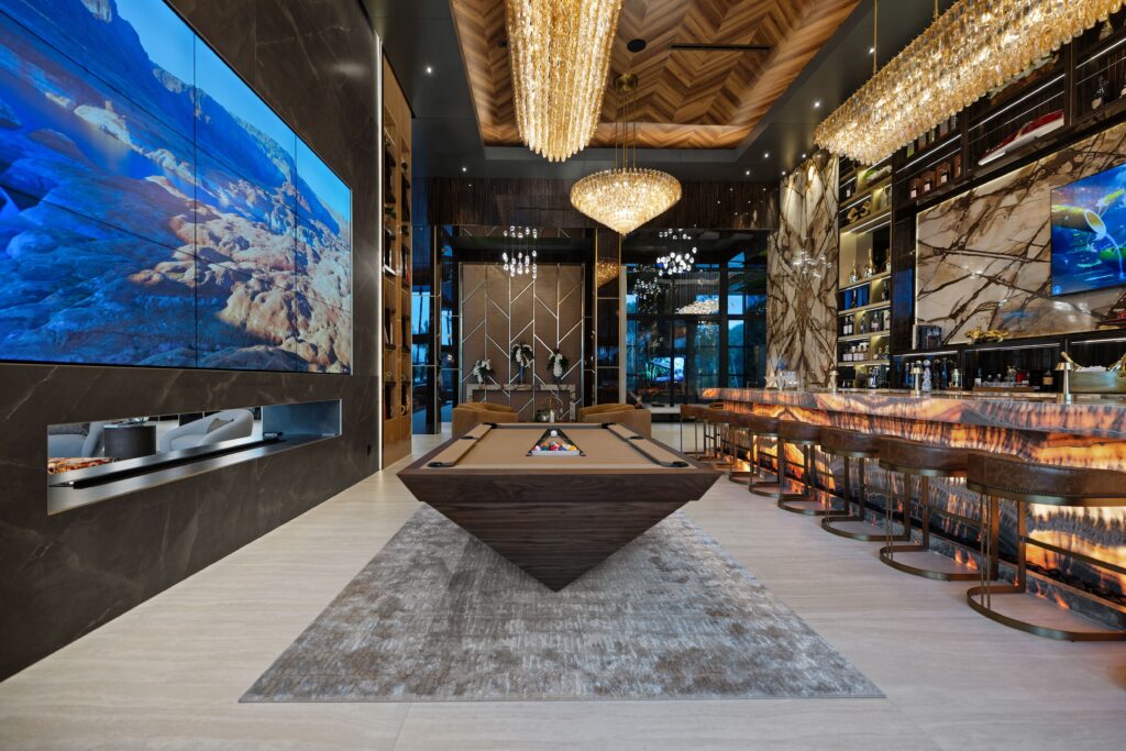 Luxurious game room featuring a large pool table by Recent Work - 11 Ravens, a modern bar with stools, elegant chandeliers, multiple large TVs on marble walls, and sophisticated decor in warm tones and wood accents.