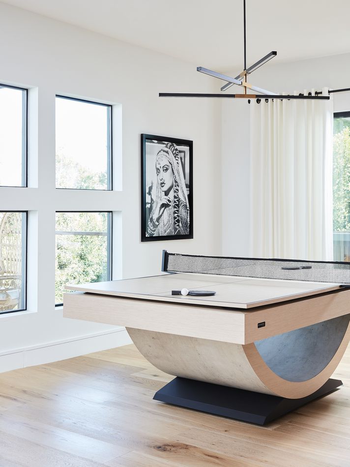Modern room with large windows, wood floors, and a stylish Recent Work - 11 Ravens ping pong table. A minimalistic black chandelier hangs overhead, while a black-and-white portrait of a woman overlooks the paddle and ball on the table.