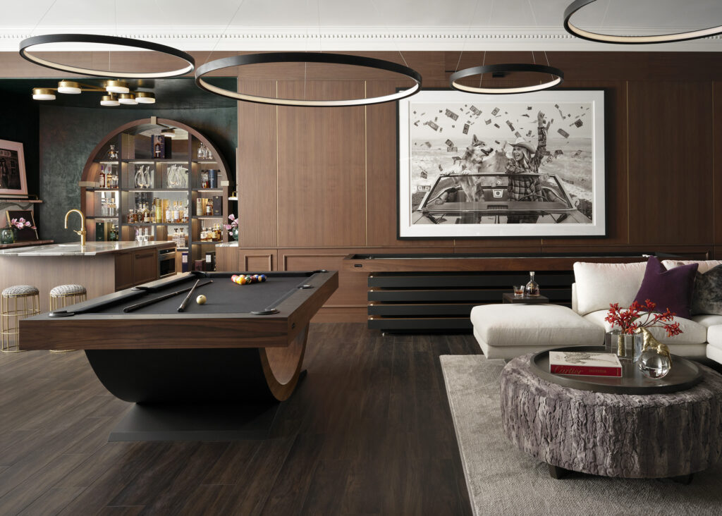 Modern game room featuring a pool table by Recent Work - 11 Ravens, a stylish bar with shelves of bottles, large black-and-white artwork on wood-paneled walls, a sectional sofa, round coffee table, and contemporary circular ceiling lights.