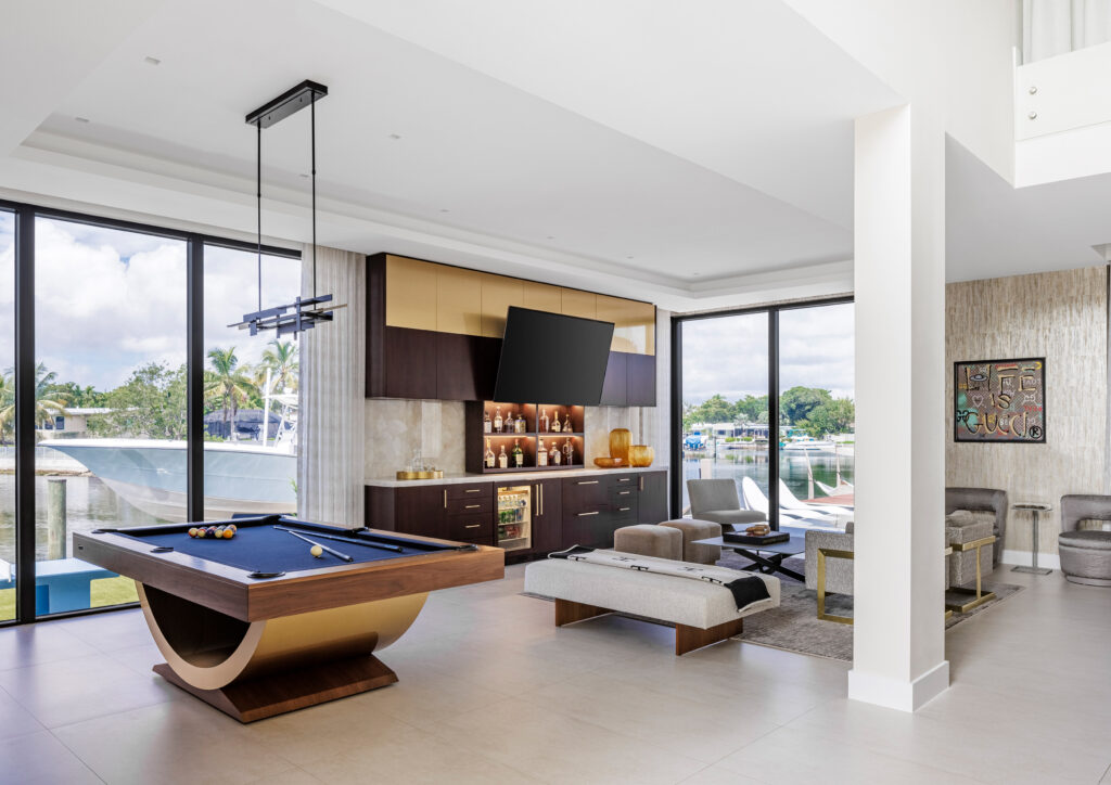 Modern living room with large windows overlooking water, featuring a contemporary 11 Ravens pool table, wall-mounted TV above a built-in bar, stylish seating, and neutral decor with natural light flooding the space. Recent Work.