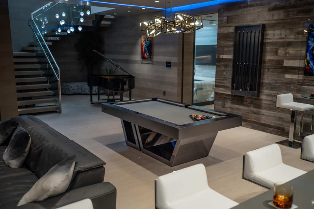 Modern game room featuring a sleek gray pool table by 11 Ravens, a rack of pool cues on a wooden wall, white bar stools, a black couch, and a staircase with glass railing. A grand piano and modern lighting complete this recent work.