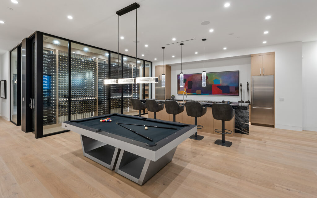 Modern room featuring a Recent Work - 11 Ravens pool table in the foreground, a sleek bar area with four black chairs, hanging pendant lights, a large wine glass display, and a colorful abstract painting on the back wall.