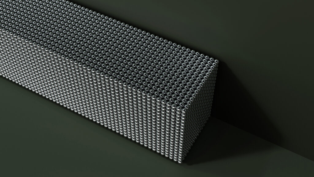 A rectangular block made up of tightly packed metallic spheres sits on a dark surface, casting a shadow. The spheres form a dense, geometric pattern across the entire block.
