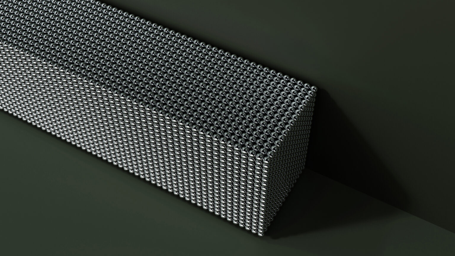 A rectangular block made up of tightly packed metallic spheres sits on a dark surface, casting a shadow. The spheres form a dense, geometric pattern across the entire block.