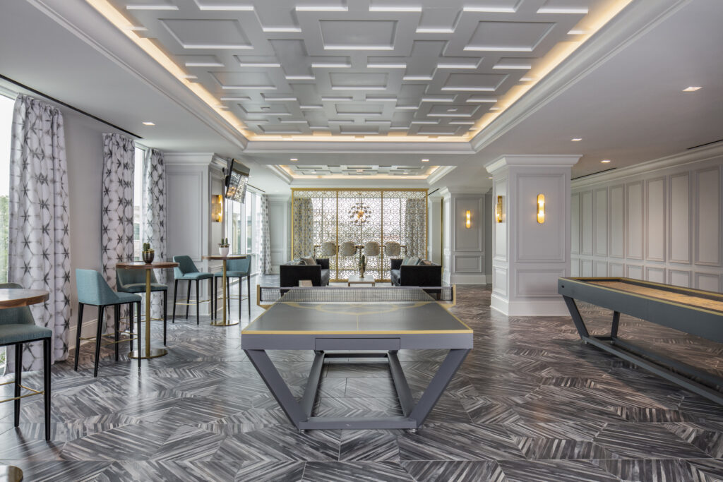 A modern game room featuring Recent Work - 11 Ravens designs, with geometric ceiling panels, patterned tile flooring, a shuffleboard table, and a ping pong table. Blue chairs and tables sit by large windows adorned with star-patterned curtains.