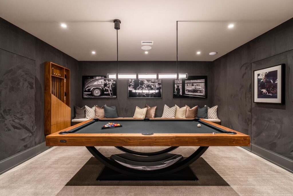 A modern game room with a black 11 Ravens pool table, cue rack, wall art, and a built-in bench with decorative pillows below framed black-and-white car photos. Recessed lighting creates a cozy, stylish atmosphere&mdash;a showcase of recent work.
